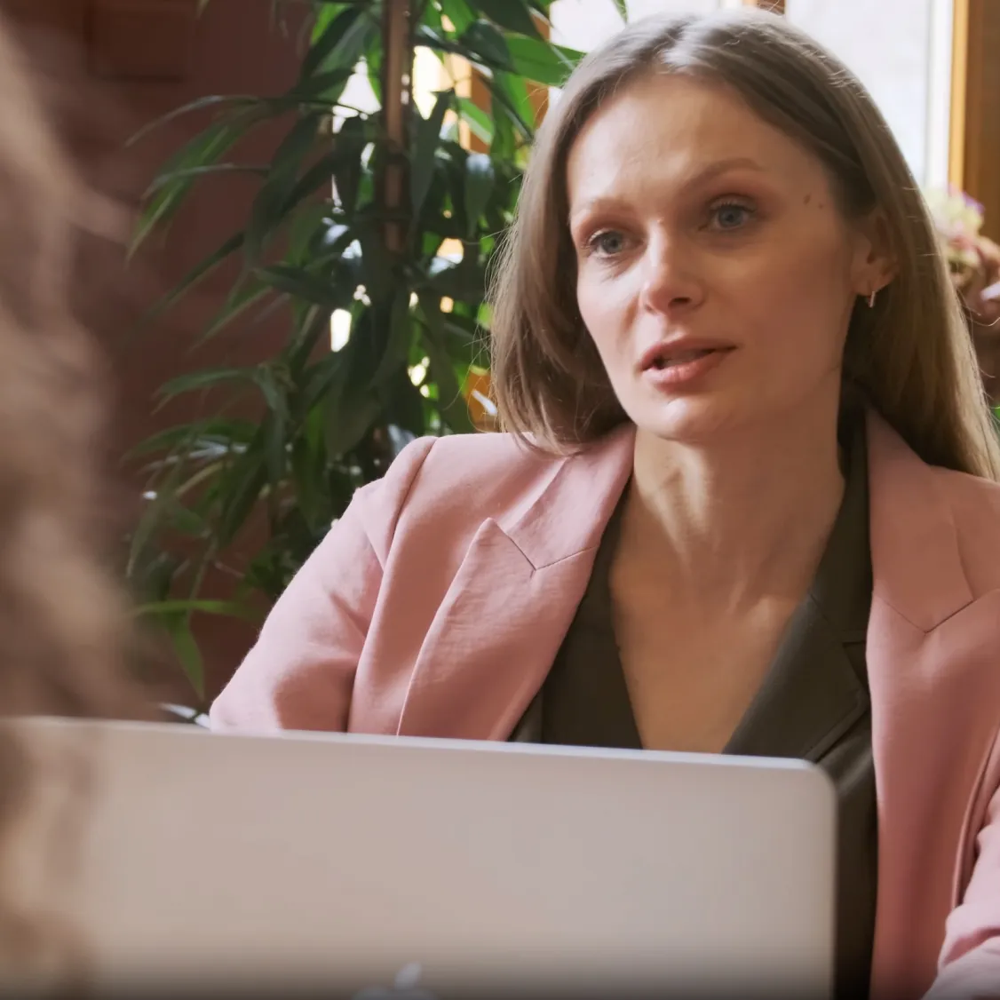 Professional woman in a pink blazer engaged in a serious conversation during a video call with a laptop in front of her.